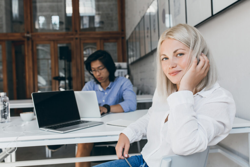 stunning female programmer playing with blonde hair while posing at workplace with asian male colleague. busy chinese web developer working with laptop sitting at the table with white secretary girl.. stunning female programmer playing with blonde hair while posing at workplace with asian male colleague. busy chinese web developer working with laptop sitting at the table with white secretary girl..